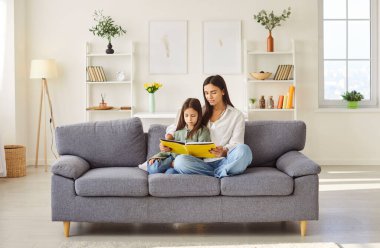 Anne ve kız kanepede kitap okuyorlar, evde mutlu vakit geçiriyorlar, anaokulu çocuğuyla birlikte evde eğleniyorlar, çocuk bakıcısı ile küçük bir kız öğrenme anlarının keyfini çıkarıyorlar, gelişiyorlar. 