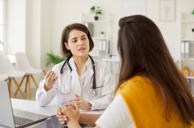 Profesyonel kadın doktor masasında oturuyor ve parlak tıp bürosunda genç bir hastaya danışıyor. Doktorun sağlık sorunlarını açıklaması, tavsiye vermesi ve randevu sırasında özen göstermesi.