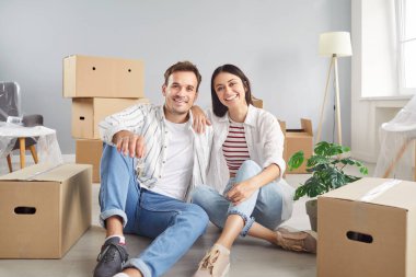 Gülümseyen ve mutlu bir aile çifti portresi. Yeni bir eve ya da daireye taşınıp kameraya bakarken yerde birlikte oturuyorlar. Neşe, sahiplik duygusu ve yeni bir başlangıç için heyecan..