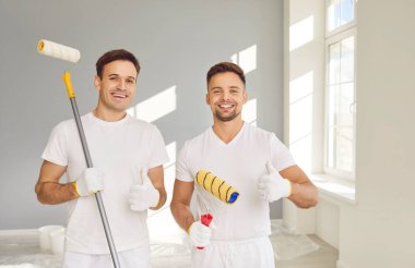 Ressamlardan oluşan gülümseyen mutlu bir ekibin portresi boya silindirleriyle kameraya bakıyor ve baş parmak işareti gösteriyor. İnşaat müteahhitleri tadilat yaparken yeni dairenin duvarını boyuyor..