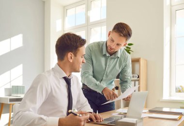 Kendine güvenen iki genç iş adamı ve şirket çalışanı ofiste dizüstü bilgisayarla çalışıyor ve iş yerindeki mali belgelere bakıyor..