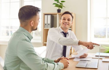 Kendine güvenen iki genç iş adamının portresi. Çalışma masasında oturmuş dizüstü bilgisayar ekranına bakıyor ve gülümsüyor. Ofisteki şirket finans verilerini tartışıyorlar..