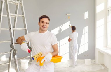 Mutlu profesyonel ressam boya silindiriyle ayakta duruyor ve arka planda duvarları boyayan bir işçiyle kameraya neşeli bakıyor. Müteahhitler tadilat yapıyor. Onarım kavramı.