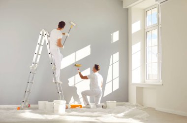Profesyonel ev yenileme servisinden iki erkek işçi duvarları açık gri renge boyuyor. Büyük, parlak bir oturma odasında. Yeni, modern bir ev ya da apartmanın içinde.