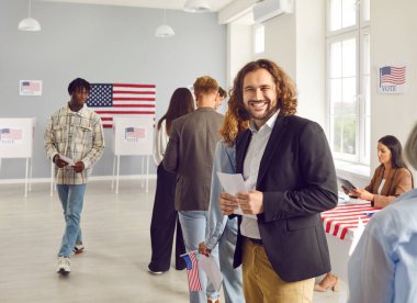 Genç mutlu Amerikalı seçmen oy merkezinde kuyrukta bekliyor ve seçim günü oy kullanma merkezindeki kameraya neşeli bir şekilde bakıyor. Seçim merkezinde oy pusulası tutan adam.