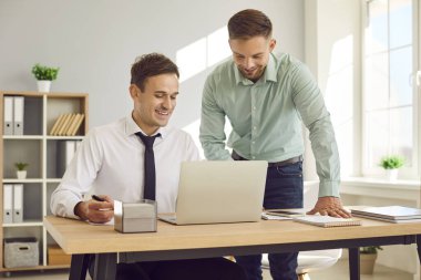 İki adam birlikte çalışıyor. Arkadaş canlısı bir adam modern ofisteki yeni meslektaşına iş danışmanlığı yapıyor. Bir adam dizüstü bilgisayarın önünde oturuyor diğeri de yanında duruyor ve ekranda onu gösteriyor..