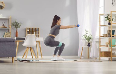 Kadın evde egzersiz yapıyor, dambıllarla çömelme hareketleri yapıyor ve spor bandı kullanıyor. Evin rahatlığında spor ve spor eğitimi rutini.