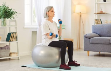 Yaşlı bir kadın spor egzersizleri yapıyor. Topun üzerine oturmuş halterleri kaldırıyor. Olgun bir kadın evdeki oturma odasında egzersiz yapıyor. Sağlıklı yaşam tarzı, fitness ve spor kavramı.