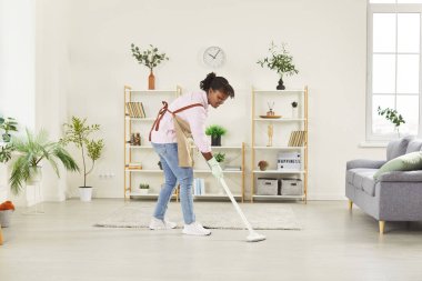 Evdeki ev işleri sırasında yerleri paspaslarken gülümseyen mutlu Afrikalı Amerikalı kadın. Ev ortamını temiz ve düzenli tutma sürecinin tadını çıkarmakla meşgul..