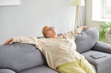 Gözlüklü ve açık renk elbiseli yaşlı bir kadın gri koltukta dinleniyor. Kolları açık ve gözleri kapalı. Yorgun bayan dinleniyor, huzurlu bir ifadeyle arkasını yaslıyor parlak, rahat oturma odasında..