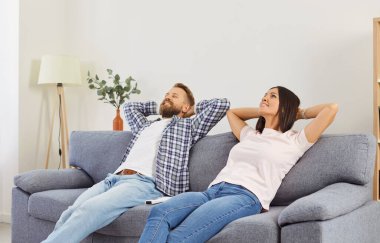 Gülümseyen genç aile çifti evdeki oturma odasında elleri kanepede otururken rahatlıyor. Karı ve koca tatmin olmuş bir şekilde dinleniyor, huzurlu bir mola veriyor..