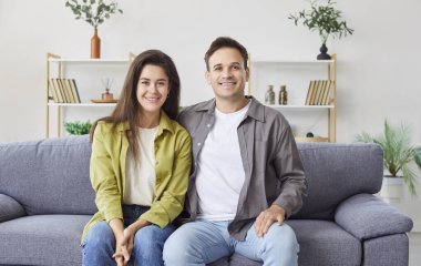 Gri koltukta yan yana oturan ve doğrudan kameraya bakan mutlu çiftin aile portresi. Evdeki kanepede kucaklaşan ve gülümseyen neşeli bir aile. Sağlıklı ahenkli ilişkiler