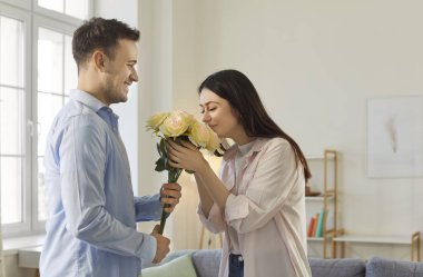 Genç adam bir buket gül çiçeğiyle tatildeki mutlu kız arkadaşını tebrik ediyor. Evdeki oturma odasında birbirine aşık, gülümseyen bir çift. Sevgililer günü ve ilişkiler konsepti.