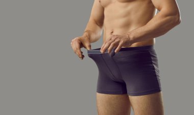 Handsome young male looks inside underpants at his private area. Shirtless attractive muscular man in underwear briefs looking at his penis standing isolated on grey background. Genital health concept