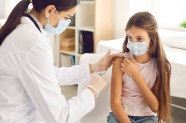 Covid-19 bağışıklık kampanyası. Yüz maskesi takan düşünceli küçük kız klinikteki doktor ya da hemşireden koronavirüse karşı aşı oluyor. Covid 19 'a karşı çocukların aşılanması kavramı.