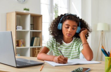 Küçük çocuk çalışmak için bilgisayar kullanıyor. Kulaklık takmış Afro-Amerikalı kız evinde dizüstü bilgisayarla oturmuş, online dersler dinliyor, elinde kalem tutuyor ve not defterine not alıyor.