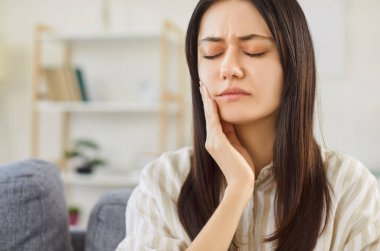 Evdeki oturma odasındaki kanepede oturan ve diş ağrısı çeken mutsuz genç bir kadının yakın plan portresi. Sıradan giysiler içindeki üzgün esmer kız diş ağrısı içinde yanağını tutuyor..