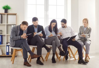 Sıraya dizilmiş sandalyelerde oturan, kağıt belgelerini tutan ve iş görüşmesine hazırlanan ya da iş görüşmesi yapan, sırada bekleyen, bazı şeyleri tartışan gençlerden oluşan bir grup.