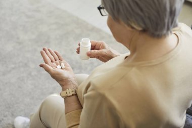 Orta yaşlı bir kadının elinde ilaç şişesi, doktor tarafından reçete edilen ilaçlar, kompleks takviyeler, vitaminler, sağlığıyla ilgilenmesi, evde olması, kronik ağrılarla başa çıkması...