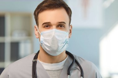 Tıbbi maskeli, steteskoplu genç bir doktorun hastaya yardım etmeye hazır kameraya bakarken, klinikte randevu verirken, tıp alanında çalışırken çekilmiş kapalı çekim görüntüleri.