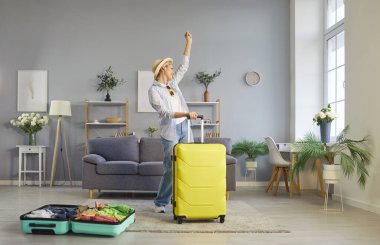 Evde bavuluyla yaz tatiline hazırlanan olumlu bir kadın dans ediyor ve kutlama yapıyor. Seyahat, seyahat planlaması ve tatil hazırlıklarının heyecanı rahat ev ortamını dolduruyor..