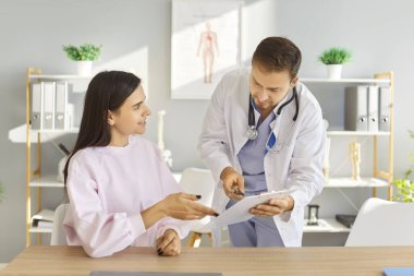 Genç bir doktor randevu ile rapor tutuyor ve tıbbi muayene sırasında klinikteki masada oturan neşeli bir bayana reçete yazıyor. Tıp konsepti.