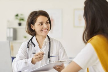 Genç gülümseyen kadın doktor klinikteki randevusunda kadın hastaya danışmanlık yapıyor, tıbbi muayene sonuçlarını açıklıyor, toplantıda tavsiye veriyor, sınav sonuçları hakkında konuşuyor, yardım ediyor, destek oluyor.