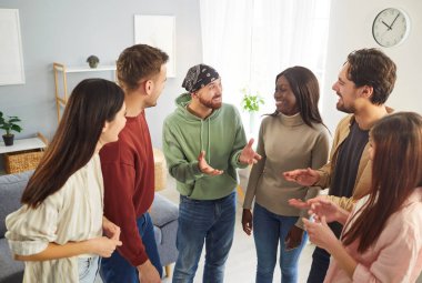 Mutlu ve çeşitli genç arkadaşlar evde çember oluşturarak konuşuyorlar. Gülümseyen erkek ve kadın öğrenciler günlük giysiler içinde birbirleriyle iletişim kurarak içeride buluşuyorlar. Arkadaşlık kavramı.