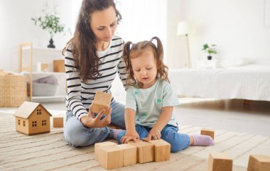 Mutlu anne ve neşeli kreş çocuğu evde halının üzerinde oynuyorlar, tahta küpleri istifleyerek, oyuncak yapı taşları inşa ederek, gülümseyerek, eğlenerek, eğlenerek, eğlenceli bebek bakıcılığı yaparak,