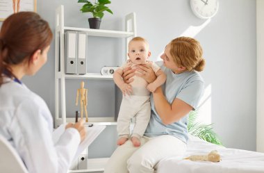Genç gülümseyen anne, tıbbi muayenede kucağında bebekle tıbbi muayenede aile doktoruna oturuyor. Bayan çocuk doktoru. İlgili anne, yeni doğmuş çocuğunu büyütüyor..