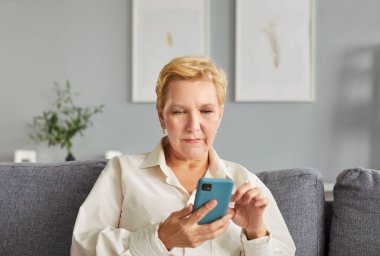 Evdeki oturma odasındaki koltukta elinde cep telefonuyla gündelik giysiler giyen kendinden emin yaşlı bir kadının portresi. Sosyal ağ internet teknolojisi kullanan yaşlı kadın.