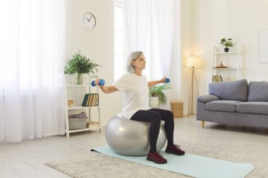 Aktif yaşlı bir kadın evde oturma odasında spor yapıyor, spor salonu ve halterlerle spor egzersizleri yapıyor, sağlık, güç ve eğitim odaklı spor yaşam tarzını benimsiyor..
