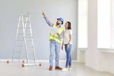 Gülümseyen genç kadın ev sahibi ustabaşıyla birlikte boş bir dairede duruyor ve bir tasarım projesi ya da onarımı tartışıyor. İç dekorasyon hakkında müteahhitle görüşeceğim. Taşınma konsepti.