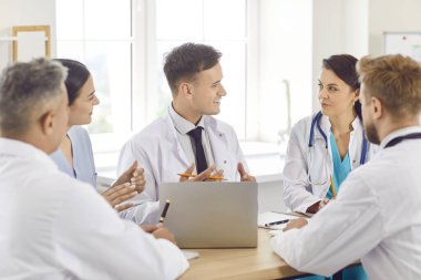 Hastanedeki ya da klinikteki iş yerinde oturup hastalara tanı koyma ya da tedavi planlama konularını tartışan kendine güvenen beyaz üniformalı genç bir ekip. İlaç ve sağlık hizmetleri kavramı.