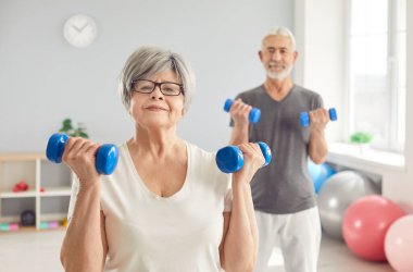 Spor egzersizleri yapan, klinikte kol rehabilitasyonu sırasında dambılları kaldıran neşeli yaşlı çiftin portresi. Neşeli gülümseyen yaşlı adam ve kadın sağlık ve sağlık için birlikte spor yapıyorlar..