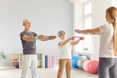 Rehabilitasyon merkezinde lastik bantla egzersiz yapan yaşlı bir çift. Eğitimci yaşlı insanlara kollarını nasıl açacaklarını öğretiyor. Fiziksel egzersiz kavramı, emekli sporcu.