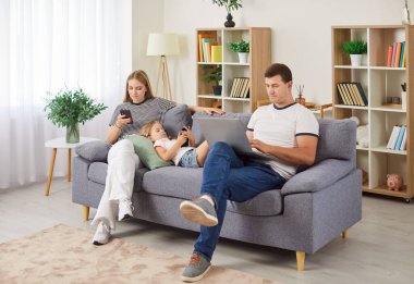 Küçük çocuklu genç ebeveynler kanepeye oturup dizüstü bilgisayar ve akıllı telefonları birlikte kullanıyorlar. Gülümseyen aile kızıyla birlikte evdeki kanepede dinleniyor. Aile ve teknoloji kavramı.