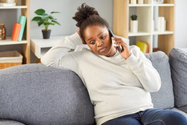 Evdeki kanepede üzgün surat ifadesiyle otururken telefonla konuşan Afrikalı Amerikalı genç bir kızın portresi. Üzgün kadın oturma odasında akıllı telefonuyla kanepede dinleniyor..