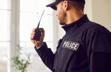 Polis telsizle konuşuyor. Emirleri iletiyor ya da görev başında çabaları koordine ediyor. POLICE yamasıyla donanmış ciddi bir polis içeride dikilirken el telsiziyle iletişim kuruyor.