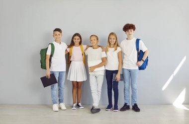 Gülümseyen mutlu okul öğrencileri kameraya neşeli bakıyorlar ve gri duvar arkasında izole edilmiş kucaklaşıyorlar. Sıraya dizilmiş sırt çantalı kızlar ve erkekler. Okul konseptine dönelim..