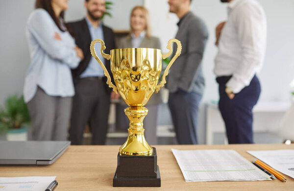 Золотой и блестящий чемпионский кубок на столе с командой деловых людей в офисе, празднующих успех, достижения и победу с наградой в профессиональной обстановке с гордостью.