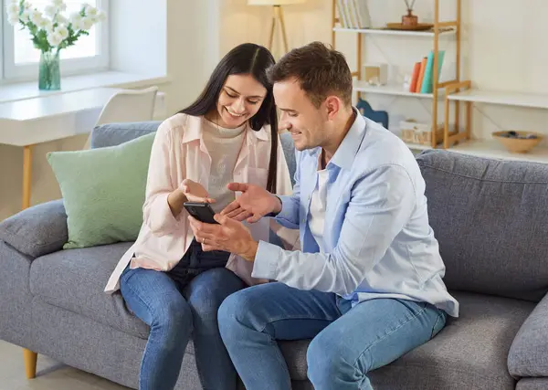Akıllı telefon kullanan neşeli genç bir çift oturma odasındaki kanepede oturmuş uygulamaları inceliyor. Genç adam gülümseyen karısına cep telefonundan video ya da haber gösteriyor. İnsanlar ve teknoloji kavramı.