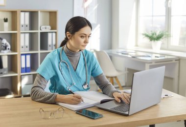 Ciddi kadın pratisyen ya da hemşire not defterine notlar yazıyor, dizüstü bilgisayarda çalışıyor, mavi üniforma giyiyor, iş yerinde kabinede poz veriyor, internetten webinar izliyor.