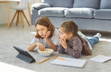 İki küçük çocuk evde halıya uzanıp dijital tablette bir şeyler izliyorlar. Odaklanmış kardeşler patlamış mısır yiyorlar ve hafta sonu boş vakitlerinde oturma odasında dinleniyorlar..