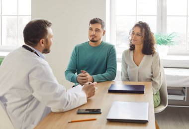 Klinikteki doktor danışman çift, tıbbi kayıtlar yazıyorlar, hasta bilgilerini dolduruyorlar, karı-kocayla konuşuyorlar, üreme sağlığı hakkında iyimser danışmanlık yapıyorlar, planlıyorlar.