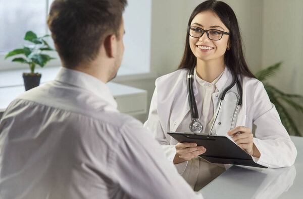 Happy young female nurse, doctor, physician woman giving healthcare consultation to male patient, explaining medical insurance benefits, illness treatment at checkup meeting, listening to help