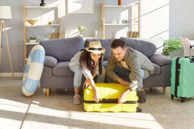 Çift, yaklaşan yaz tatiline hazırlanmak için bavullarını hevesle topluyor. Gündelik giysiler içinde kadın ve erkek, evde yerde otururken fermuarını kapatmaya çalışmak komiktir..