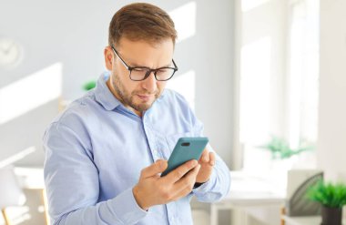 Gözlüklü, parlak bir odada ya da ofiste duran akıllı telefon ekranını ciddi bir ifadeyle yakından inceleyen odaklanmış bir adamın portresi. Yetişkin erkek, cihazında önemli bir şeyi analiz ediyor ya da okuyor..