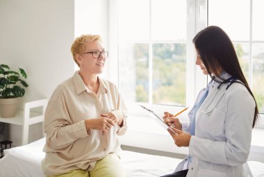 Klinikteki sağlık muayenesi sırasında elinde randevu ve danışmanlık ile rapor tutan bir kadın doktorla konuşan yaşlı bir hastanın portresi.