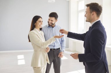Profesyonel emlakçı, ortağı yanında durup mutlu bir şekilde izlerken boş bir odada gülümseyen genç bir kadının anahtarlarını veriyor. Ev anahtarlarının alınmasını kutlayan aile çifti.